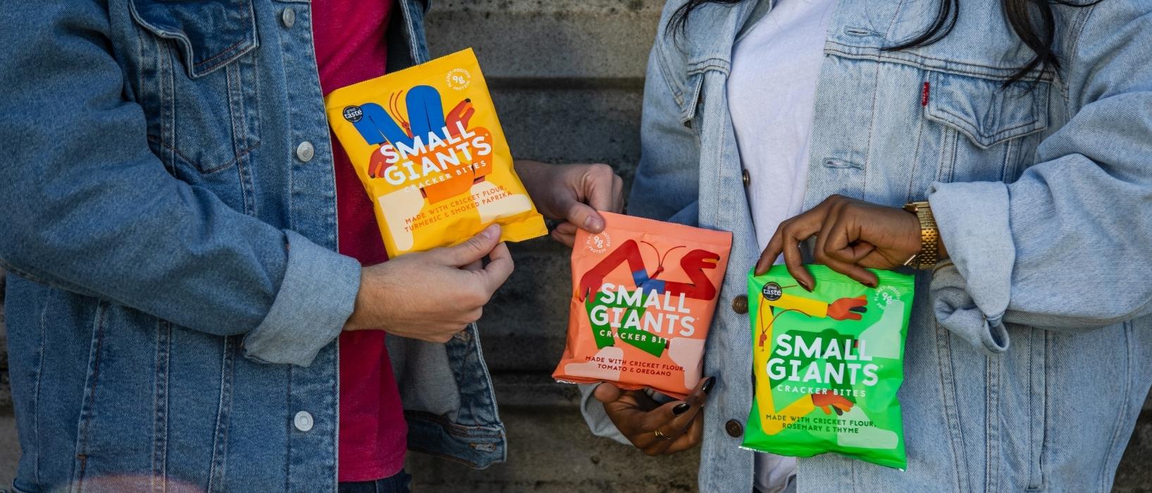 two people shot so you can just see their hands and body, they are holding three brightly coloured crisp packets with the words Small Giants on them