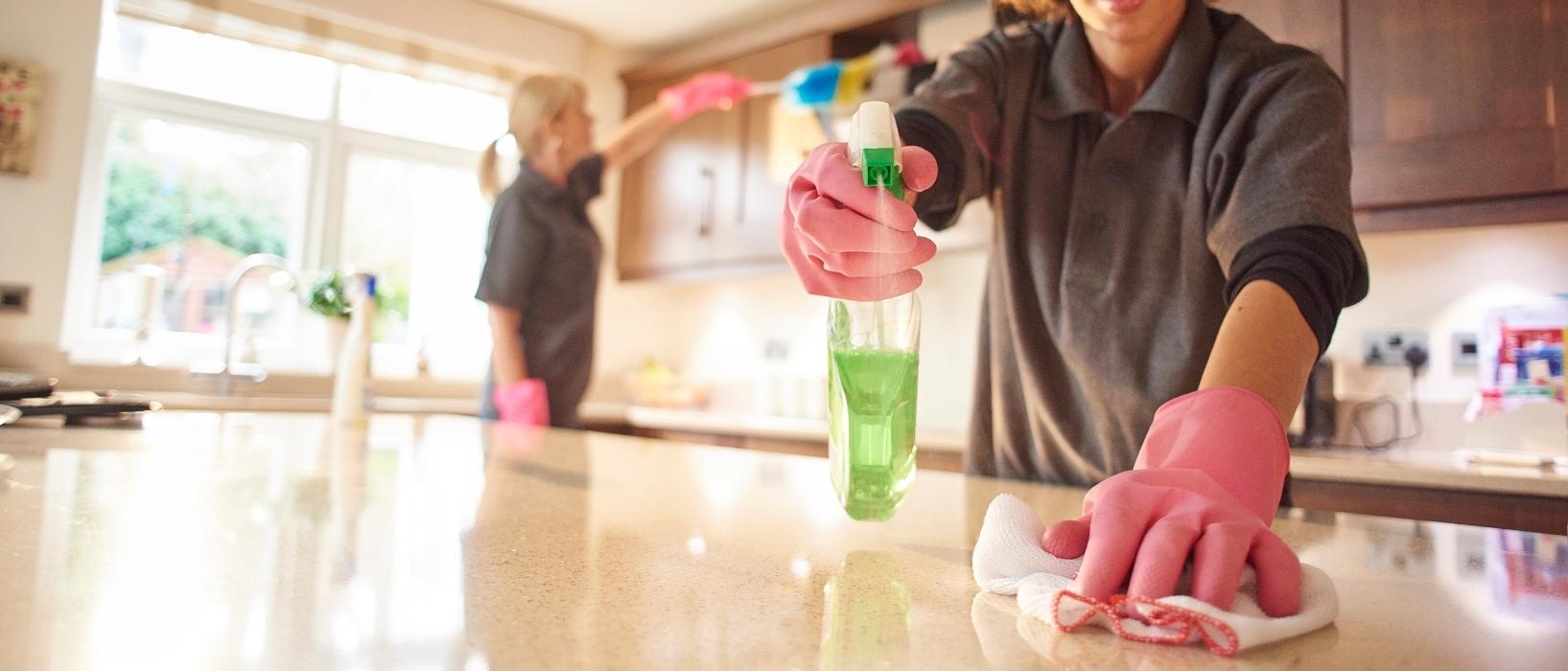 close up of woman's gloved hand and spray bottle cleaning kitchen counter, another woman is blurred in the background cleaning an oven