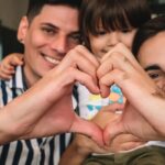 close up of two fathers with small child sitting between their heads on their shoulders, both are holding one hand up towards the camera and cupping them together to form a heart in front of child