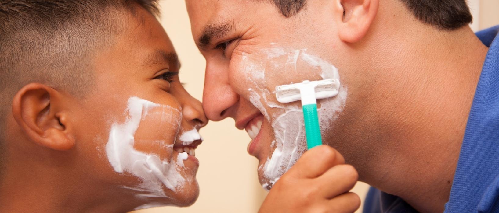 close up of rather and sons faces, noses touching and they are both smiling, shaving cream on their faces. Son is holding up a sustainable razor to his fathers cheek