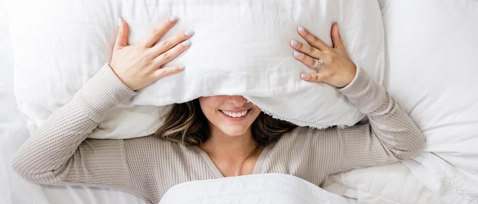 woman lying in bed with white sheets with pillow over her face so you can only see her arms and month smiling